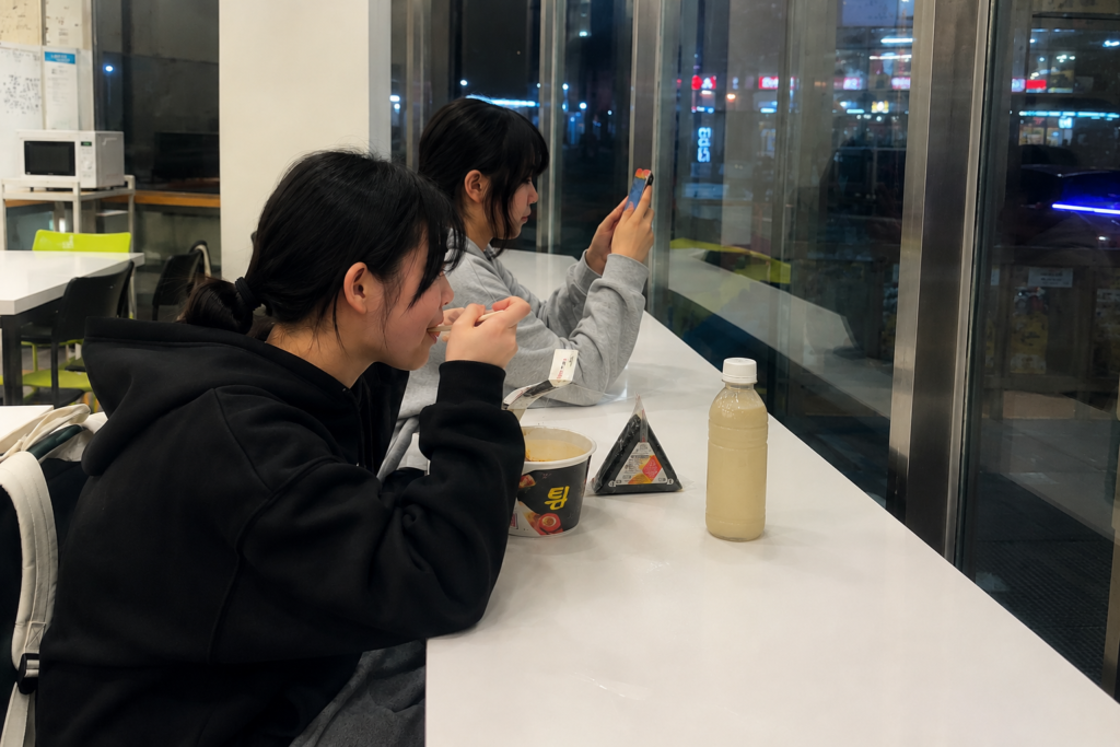 Why Do Koreans Visit Convenience Stores So Often? Korean students eating instant ramen and triangle kimbap at a convenience store seating area.